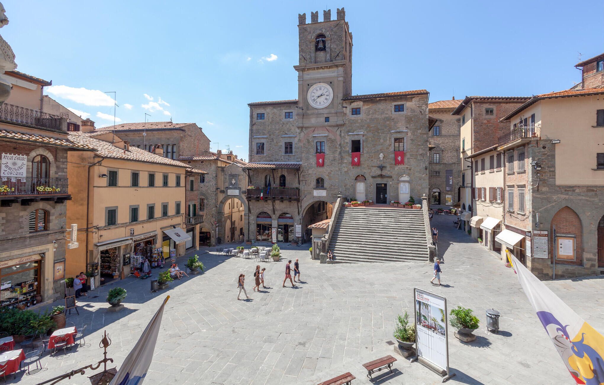 A historic square with a prominent clock tower, stone buildings, and outdoor seating areas.