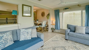 A living room with a sofa, a dining table with chairs, a ceiling fan, and a window with curtains.