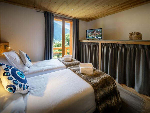 A bedroom with two beds, a wooden ceiling, a window with curtains, a television on a shelf, and a view of mountains.