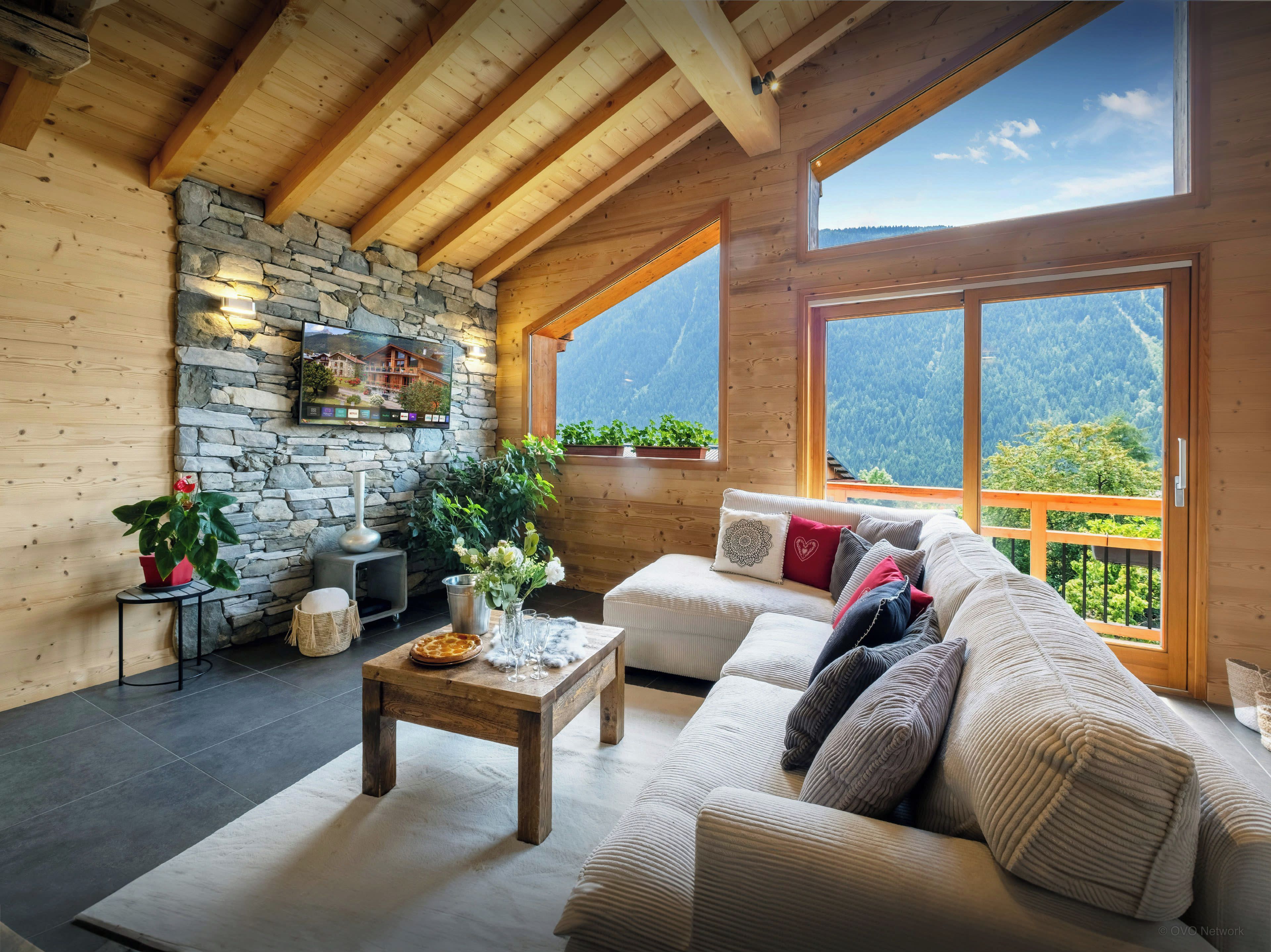 A cozy room with a stone wall, wooden beams, a sofa, a coffee table, and a view of mountains.