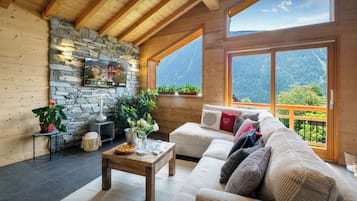 A cozy room with a stone wall, wooden beams, a sofa, a coffee table, and a view of mountains.