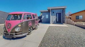 Ein pinkfarbener VW-Bus ist vor einem modernen Gebäude mit einer blauen Tür und einem kleinen Fenster geparkt.