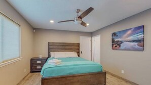 Een slaapkamer met een bed, een plafondventilator, een nachtkastje, een schilderij en een raam met jaloezieën.