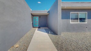 Een modern gebouw met een betonnen gevel, een smal pad dat leidt naar een turquoise deur, en een raam aan de rechterkant.