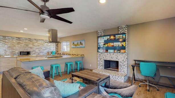 Een moderne woonkamer met een leren bank, een houten salontafel en teal accentstoelen. Een open haard met een versierd omlijsting is zichtbaar, evenals een keukenhoek met een magnetron en barstoelen.