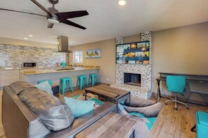 Una sala moderna con un sofá de cuero, una mesa de centro de madera y sillas de acento color turquesa. Se observa una chimenea con un marco decorativo, junto con una zona de cocina que incluye un microondas y taburetes de bar.