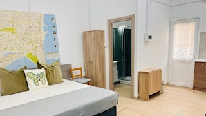 A bedroom with a bed, a map on the wall, a wooden wardrobe, a bathroom with a glass door, and a small wooden table. - Larnaca - Studio - Sleeps 4 - Garden - Parking (Larnaca)