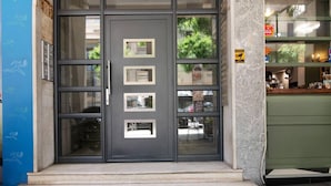 A building entrance with a dark grey door, multiple windows, and a small outdoor seating area visible through the glass. - Beautiful View 8floor Loft Apartment in Athina (Athens)