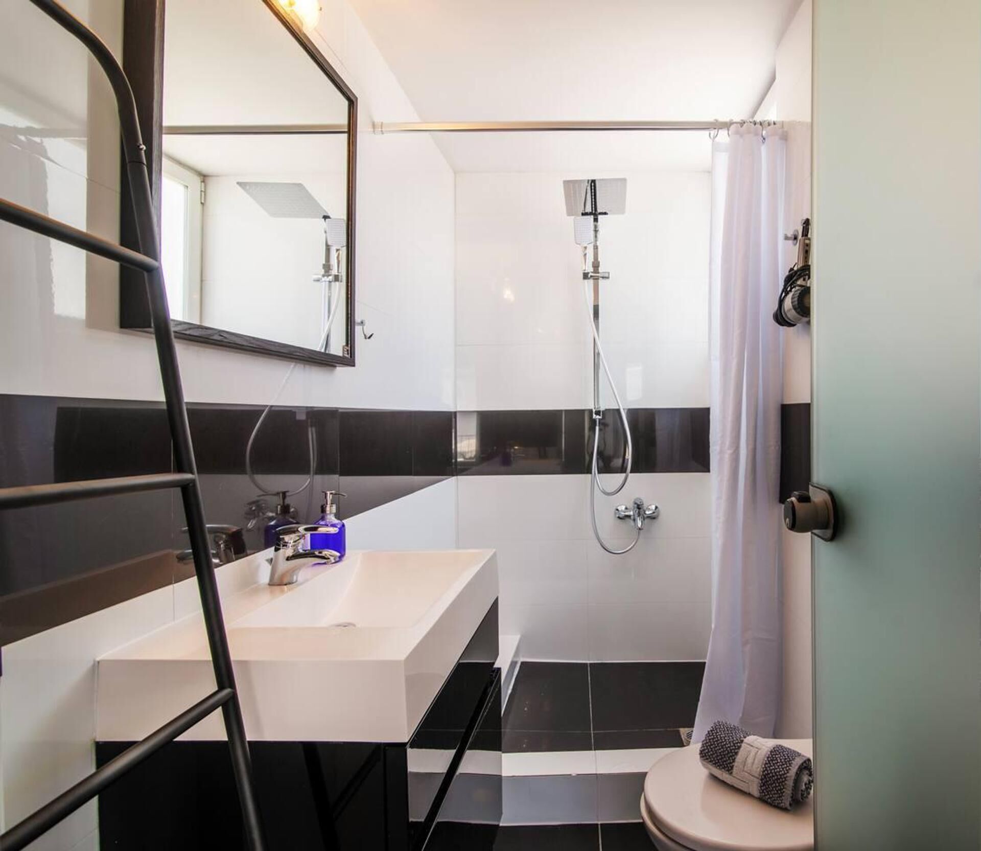 Une salle de bain moderne avec un lavabo blanc, des murs carrelés en noir et blanc, une douche avec un pommeau de douche à main et un porte-serviettes.