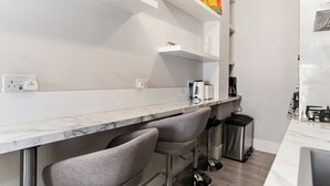 A modern kitchen with a marble countertop, two bar stools, a coffee machine, and a trash bin. - GuestReady - Modern Studio | Kensington Gem (London)