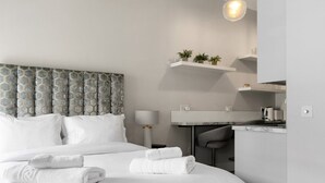 A modern hotel room with a large bed, a desk with a chair, a small kitchen area, and a ceiling light. - GuestReady - Modern Studio | Kensington Gem (London)