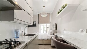 A modern kitchen with a gas stove, sink, and marble countertops. There is a dining area with a table and chairs, and a large window providing natural light. - GuestReady - Modern Studio | Kensington Gem (London)