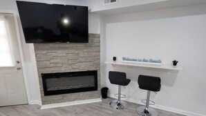 A modern living room with a flat-screen TV mounted on the wall, a fireplace, two black bar stools, and a shelf with a 'Welcome to the Beach' sign.