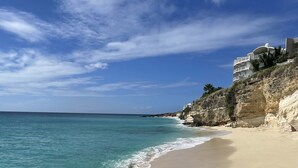 A beach with clear turquoise water, a sandy shore, and a cliff with buildings on top.