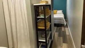 Un couloir étroit avec un plancher en bois, une baignoire blanche sur la gauche, une étagère noire avec des serviettes pliées, et un lit avec des oreillers jaunes à l’arrière-plan.
