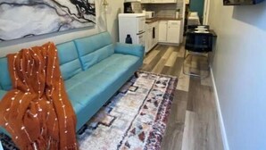 Un espace de vie compact comprenant un canapé, un tapis à motifs, un coin cuisine et un téléviseur fixé au mur.