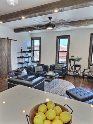 A modern living room with a large sectional sofa, a dining table with a bowl of lemons, and a ceiling fan.