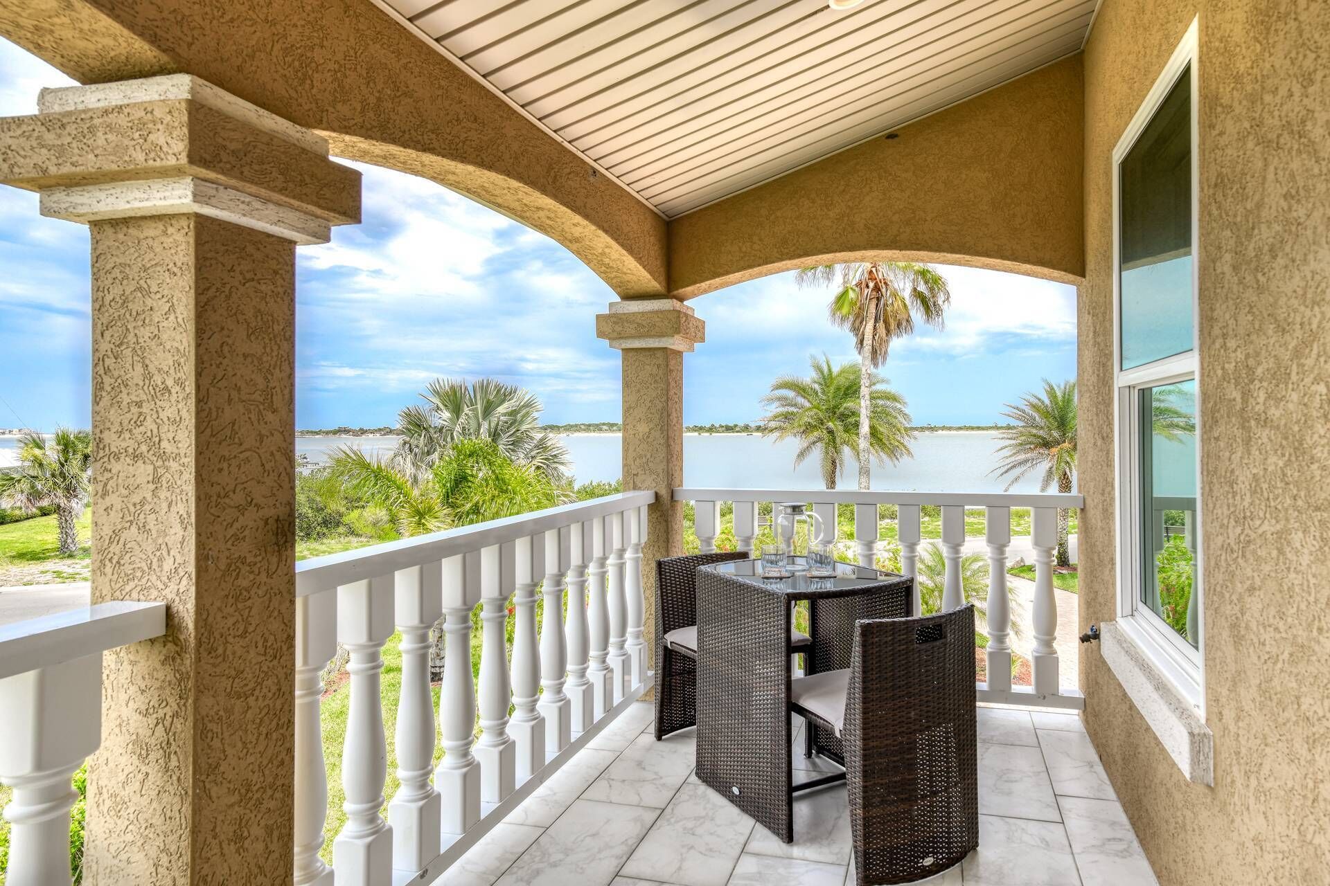 A covered balcony with a table and chairs, overlooking a body of water and palm trees.