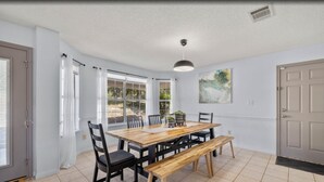 Un coin repas avec une table en bois, des chaises noires et un banc. De grandes fenêtres sont présentes, ornées de rideaux blancs, ainsi qu’une bouche d’aération au plafond et une porte sur le côté droit.
