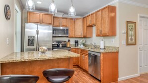 A modern kitchen with wooden cabinets, stainless steel appliances, a granite countertop, and a bar area with two stools. - Anastasia Suite (St. Augustine)