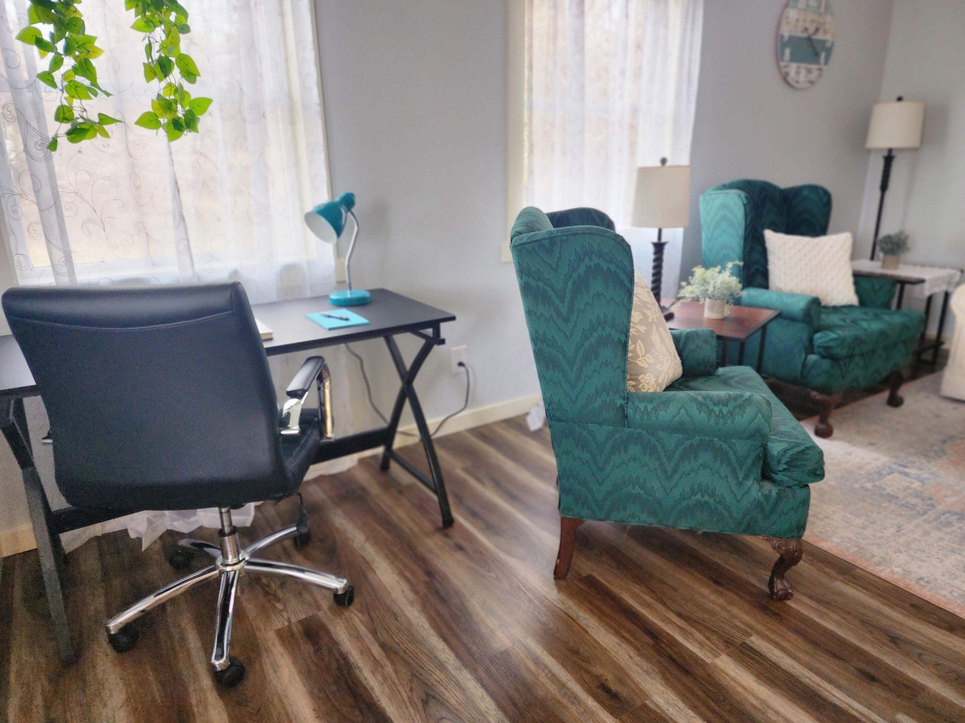Uma sala de estar moderna com uma cadeira de escritório preta, um poltrona azul-turquesa e um sofá combinando. Uma pequena mesa com um abajur e um objeto decorativo está posicionada no canto.