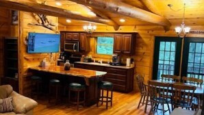 Interno di una baita in legno con un tavolo da pranzo, sgabelli da bar, un divano e un angolo cottura con televisore.