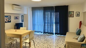 Een kamer met een eettafel, stoelen, een bank, een groot raam met gordijnen en kunstwerken aan de muur.