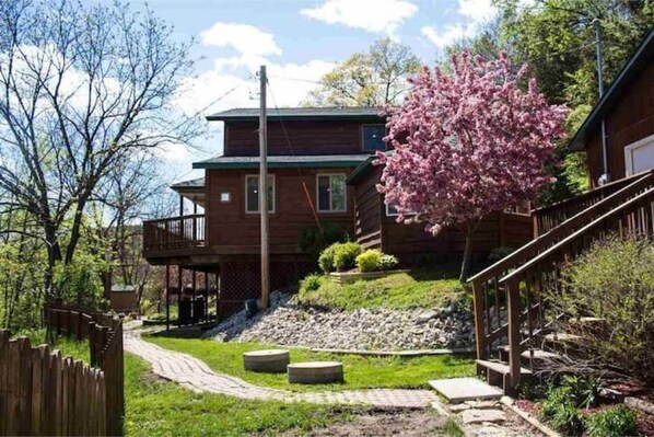 A wooden cabin with a deck, surrounded by a garden with a stone pathway and a cherry blossom tree. - Drop Tine Ridge w/Hot Tub (Lansing)