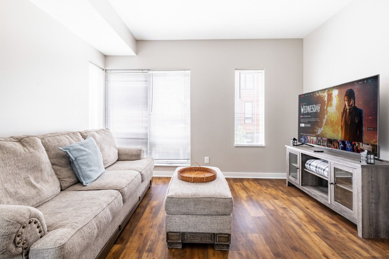 A living room with a beige sofa, a grey ottoman, a wooden TV stand, and a flat-screen TV displaying a movie.
