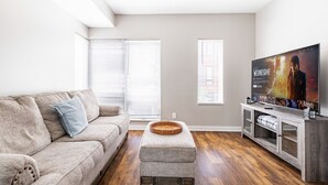 Ein Wohnzimmer mit einem beigefarbenen Sofa, einem grauen Hocker, einer Holz-TV-Konsole und einem Flachbildfernseher, der einen Film zeigt.