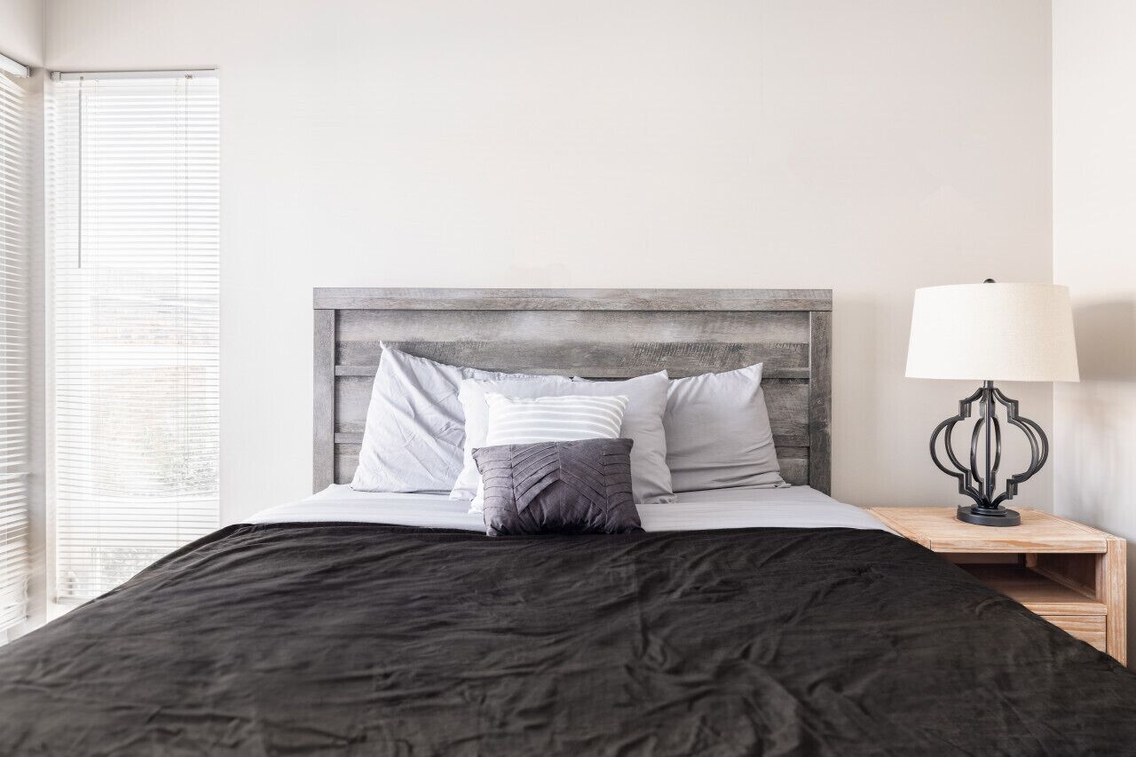 Un lit bien fait, recouvert d’une couvre-lit sombre, avec une tête de lit en bois et une lampe de chevet posée sur une table de chevet en bois.