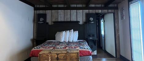 A loft-style bedroom with a wooden ceiling, a large bed with a quilt, a chest of drawers, and a hanging light fixture.