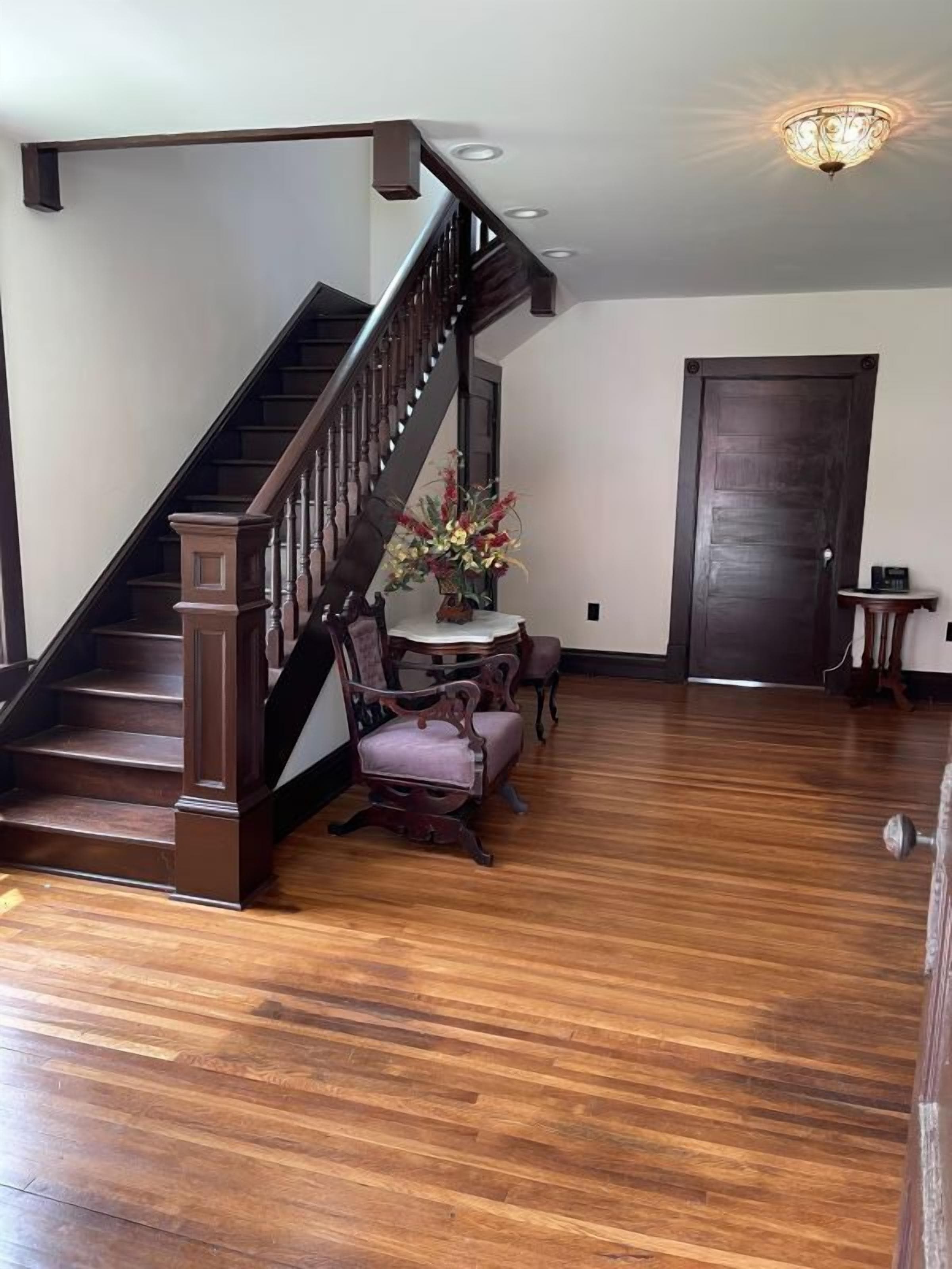 A spacious living area with a wooden staircase, a dining table with chairs, and a dark wooden door.