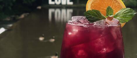 Un vaso de bebida roja con hielo, adornado con una rodaja de naranja y hojas de menta, frente a un cuerpo de agua con patos y un cartel que dice "LOVE".