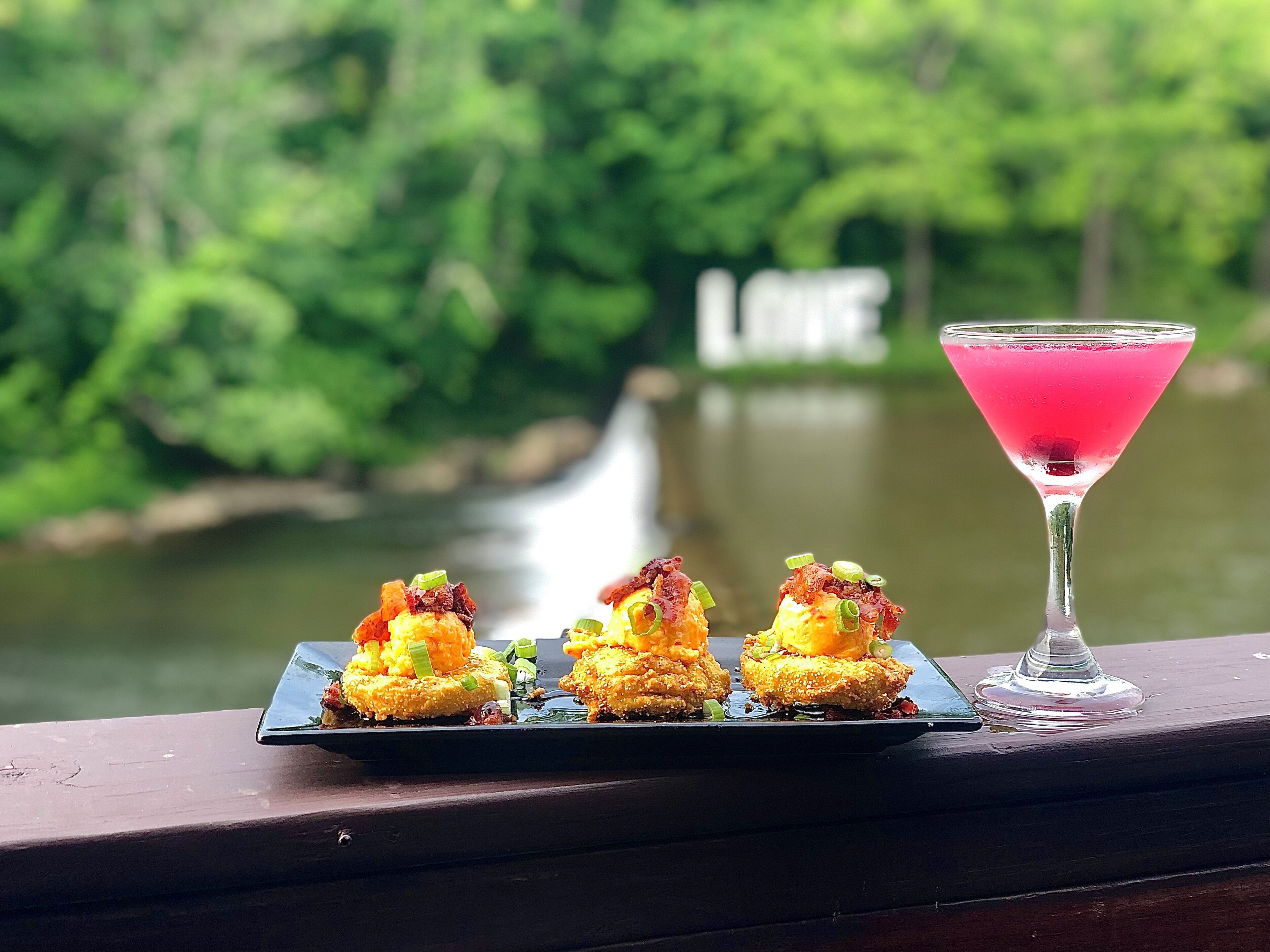A table with three appetizers and a pink cocktail on a wooden railing with a river and greenery in the background.