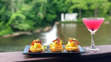 A table with three appetizers and a pink cocktail on a wooden railing with a river and greenery in the background.
