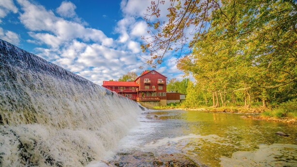 Ein Wasserfall fließt in einen Fluss, im Hintergrund steht ein rotes Gebäude.