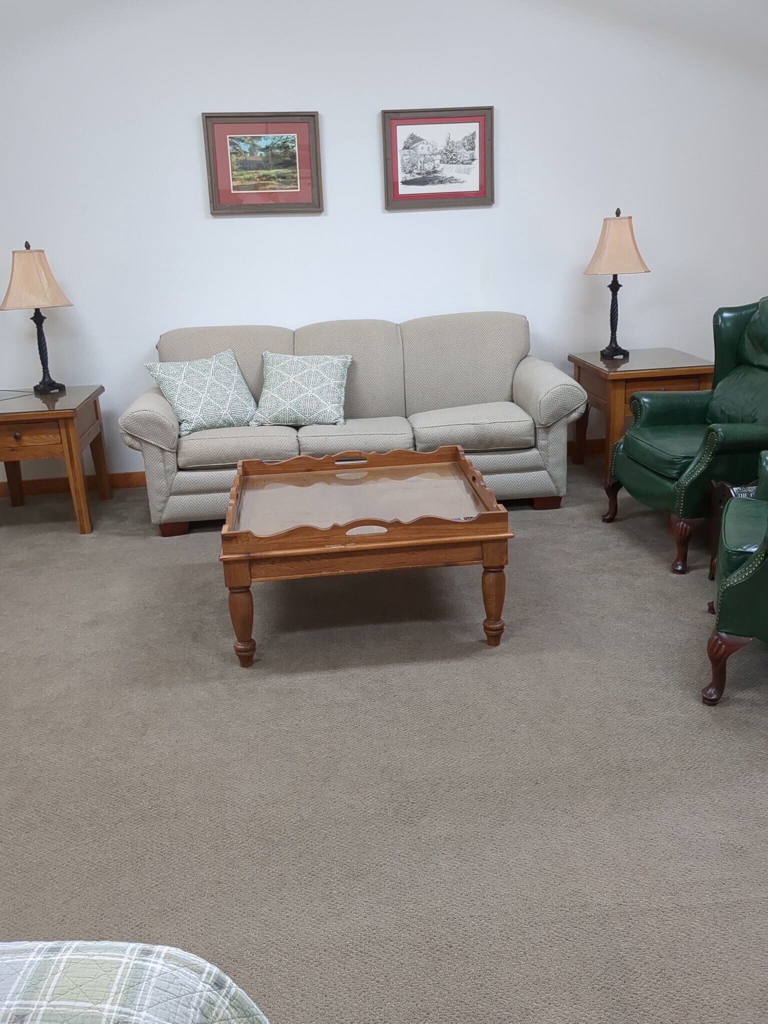 A living room with a beige sofa, two framed pictures on the wall, a wooden coffee table, a green armchair, and two side tables with lamps.