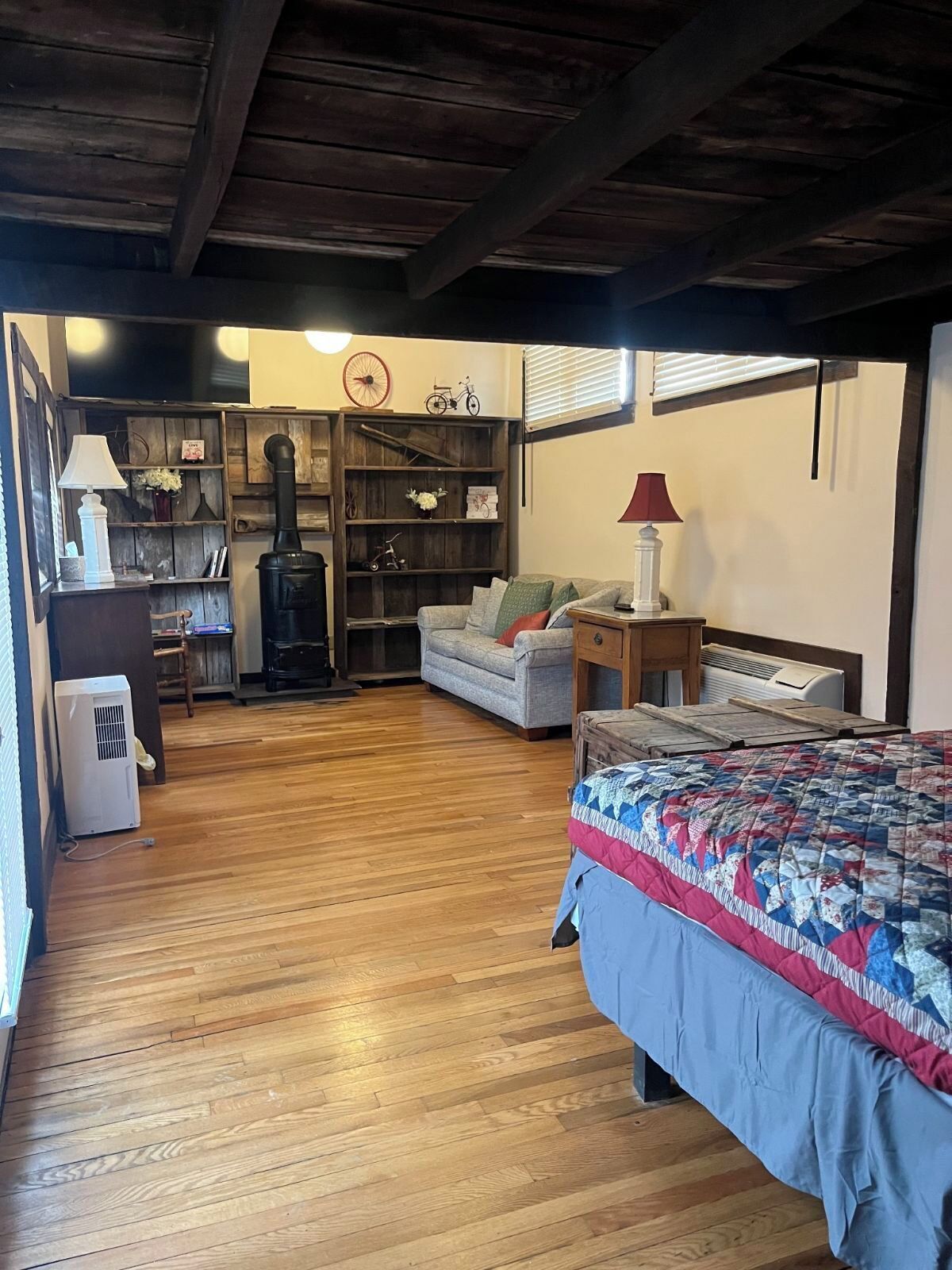 Une pièce chaleureuse avec un plancher en bois, un lit recouvert d’une courtepointe, un canapé, une petite table avec une lampe, un radiateur et une bibliothèque remplie de livres et d’objets décoratifs.