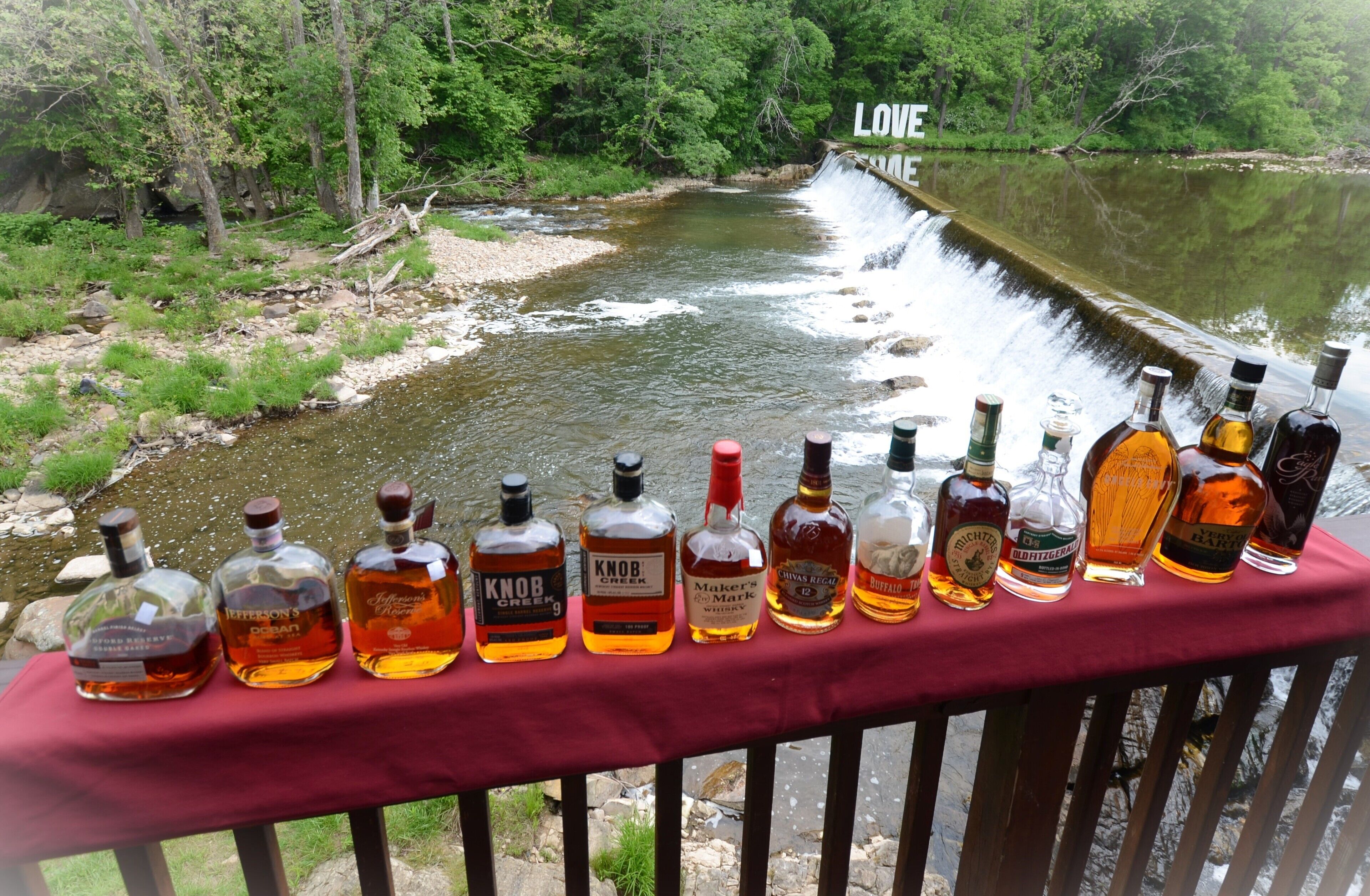 Une rangée de bouteilles de whisky sur une table rouge, près d’une rivière, avec une cascade en arrière-plan.