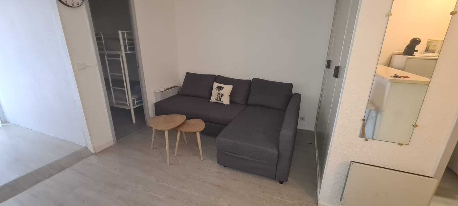 A small room with a grey sofa, two small wooden stools, a white wall clock, and a white cabinet with a mirror.
