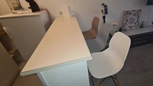A room with a white desk, a white chair, a small table with a lamp, a wall-mounted TV, and a wall decoration.
