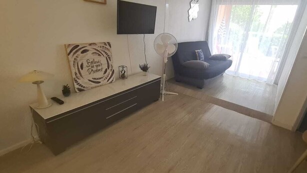 A living room with a dark sofa, a flat-screen TV mounted on the wall, a small table with a lamp, and a decorative sign.