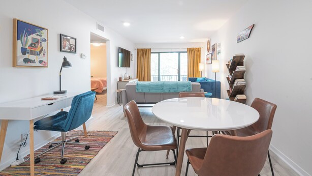 Un salon moderne avec une table à manger blanche, des chaises bleues et marron, un canapé gris et une bibliothèque.