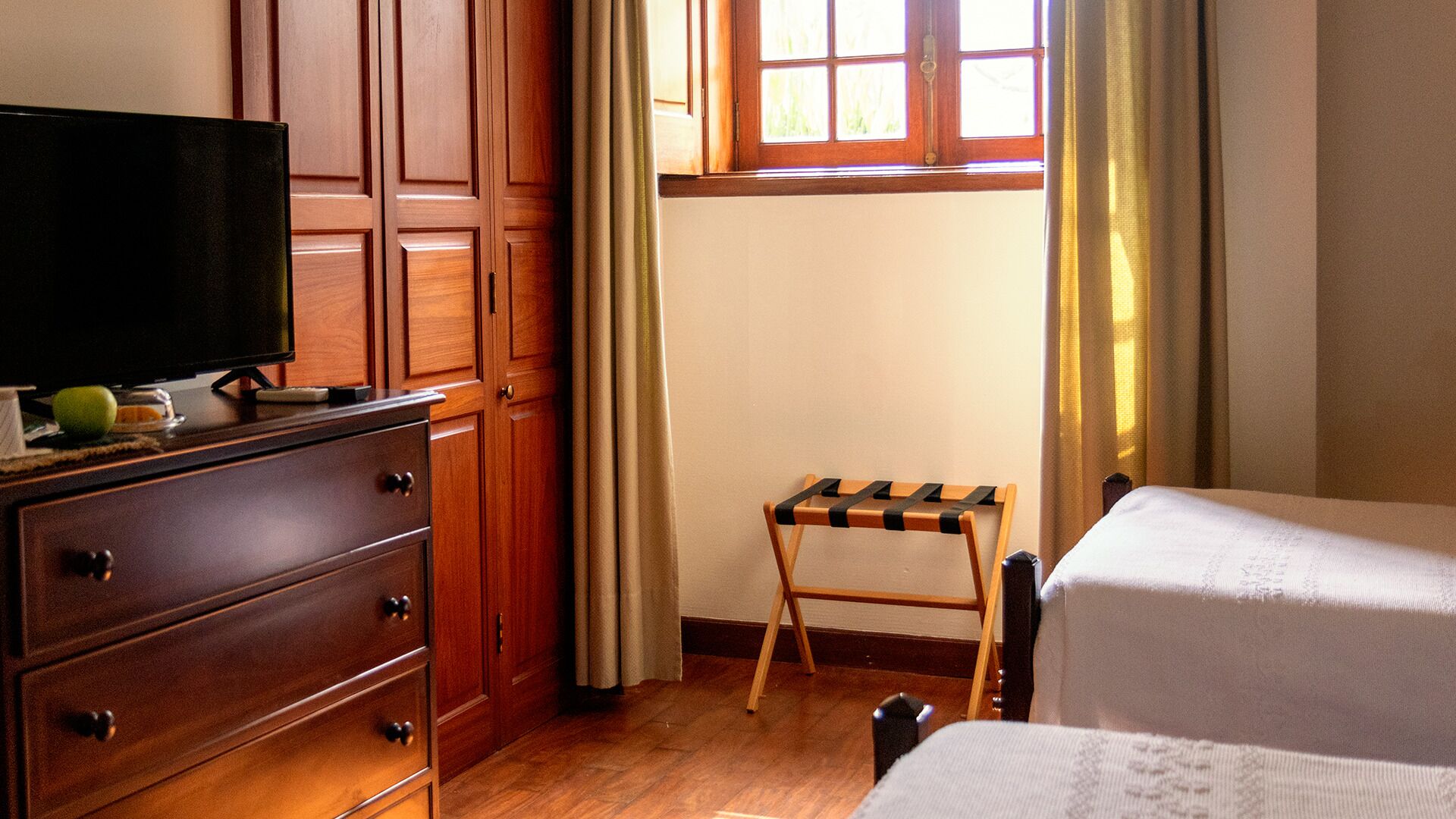 A hotel room with wooden furniture, a television on a dresser, a small table, and a window with curtains.