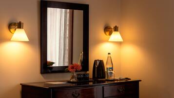 A hotel room with a dark wooden dresser, a mirror, two wall-mounted lamps, a vase with flowers, a bottle, and two black containers labeled "VANI VA".