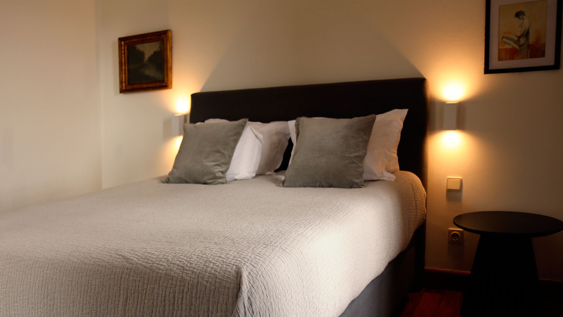 A neatly made bed with a textured white duvet, two grey pillows, and a dark headboard. There are wall-mounted lights on either side of the bed, and a small round table with a lamp to the right.