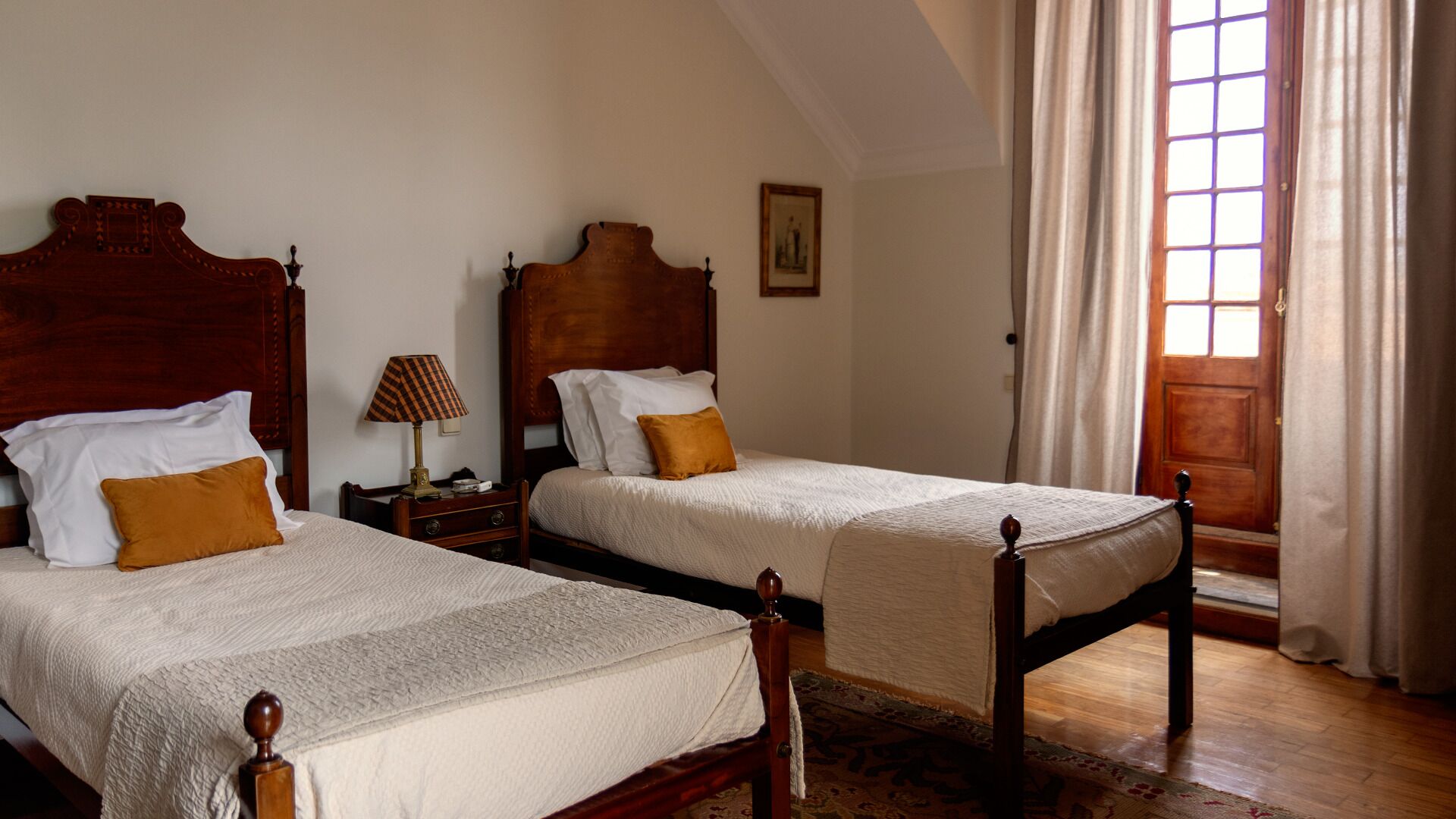Deux lits en bois, recouverts de draps blancs et agrémentés d’oreillers marron, se trouvent dans une pièce au plancher en bois et dotée d’une fenêtre avec des rideaux.