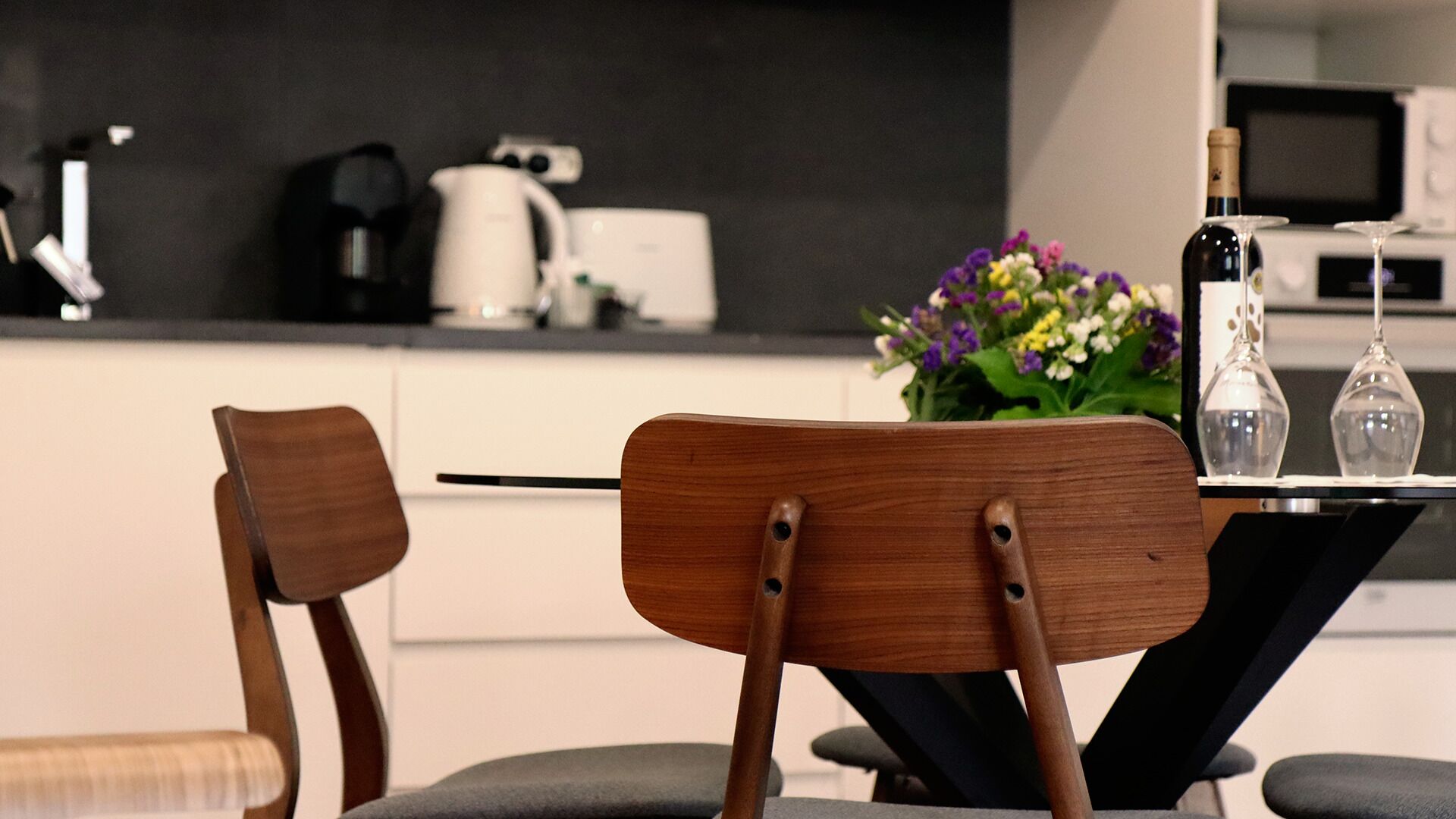 A modern kitchen with two wooden chairs, a dining table, a flower arrangement, and a bottle of wine.