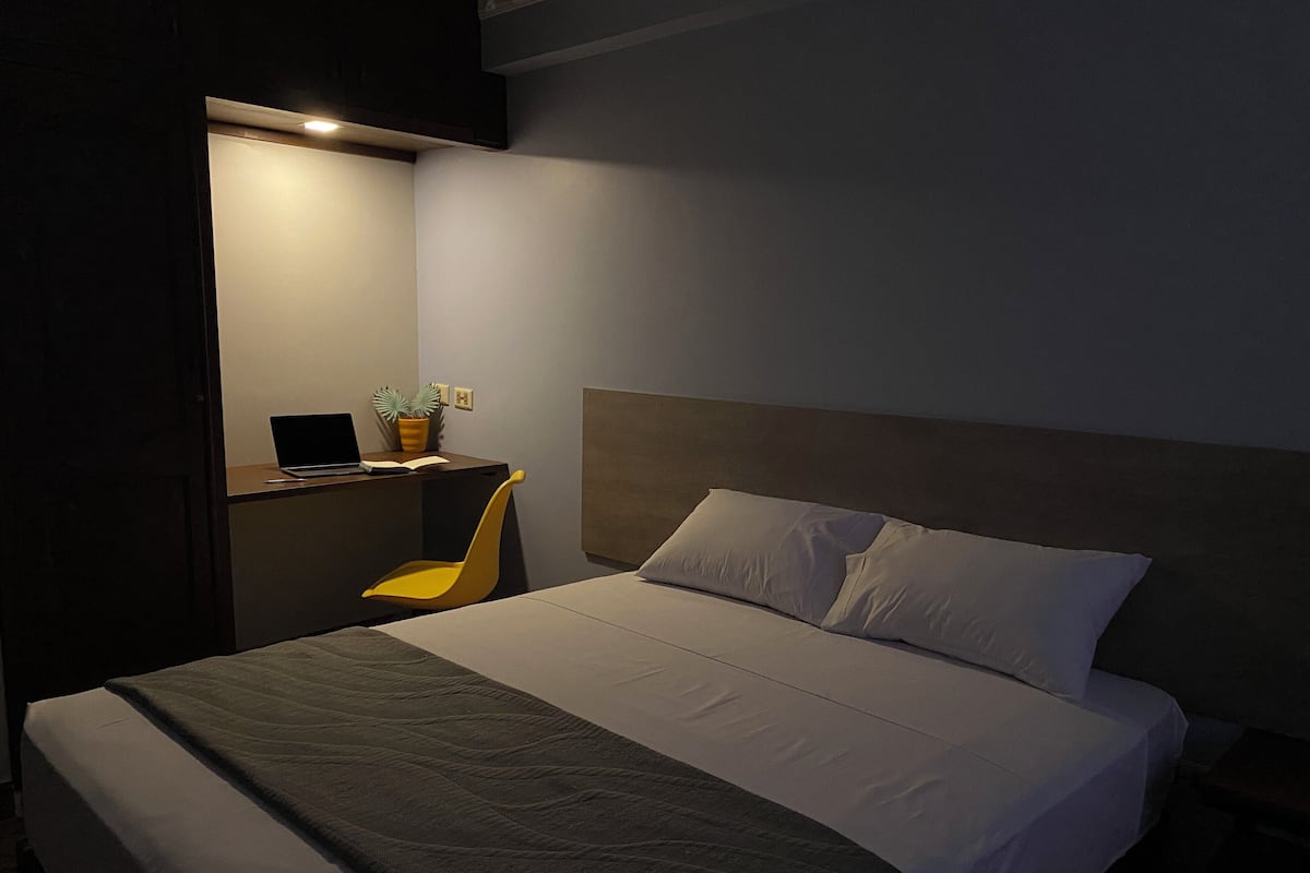 Una cama bien hecha con una manta gris, un cabecero de madera y un pequeño escritorio con una computadora portátil y una planta en maceta.